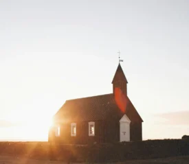 Small dark wooden church standing in an open landscape at sunrise, with warm sunlight shining behind the building and a clear sky above