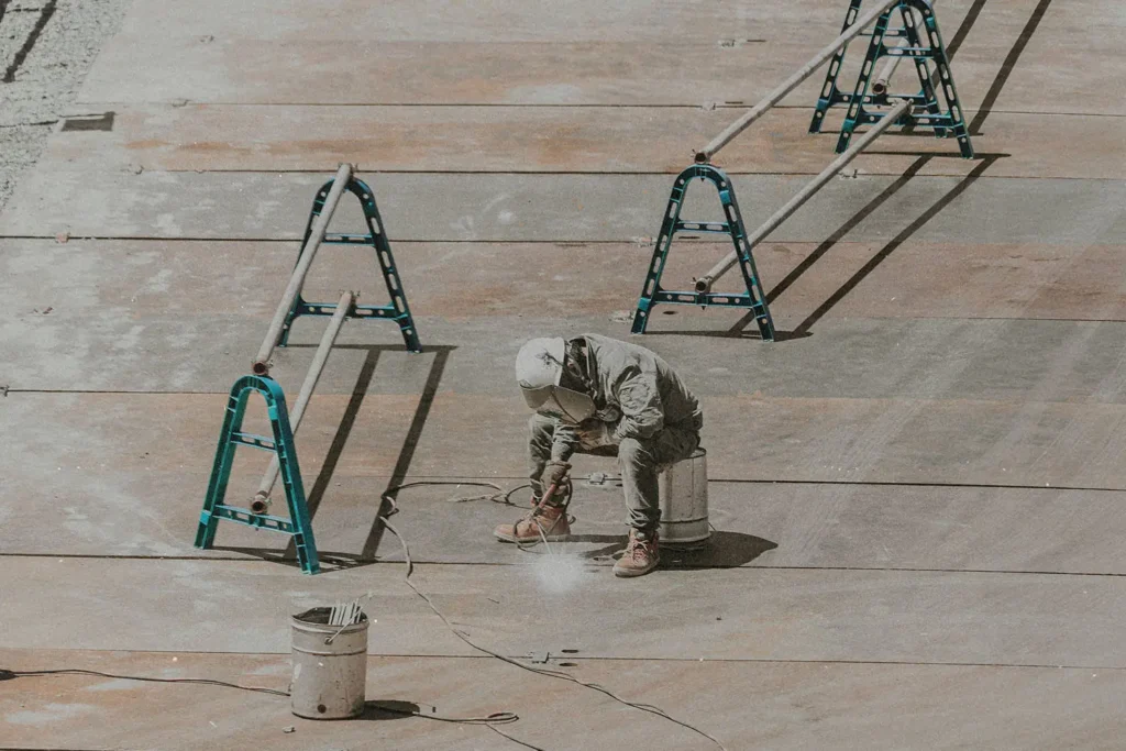 A construction worker wearing protective gear crouches on a large metal surface while using a tool that emits sparks or dust