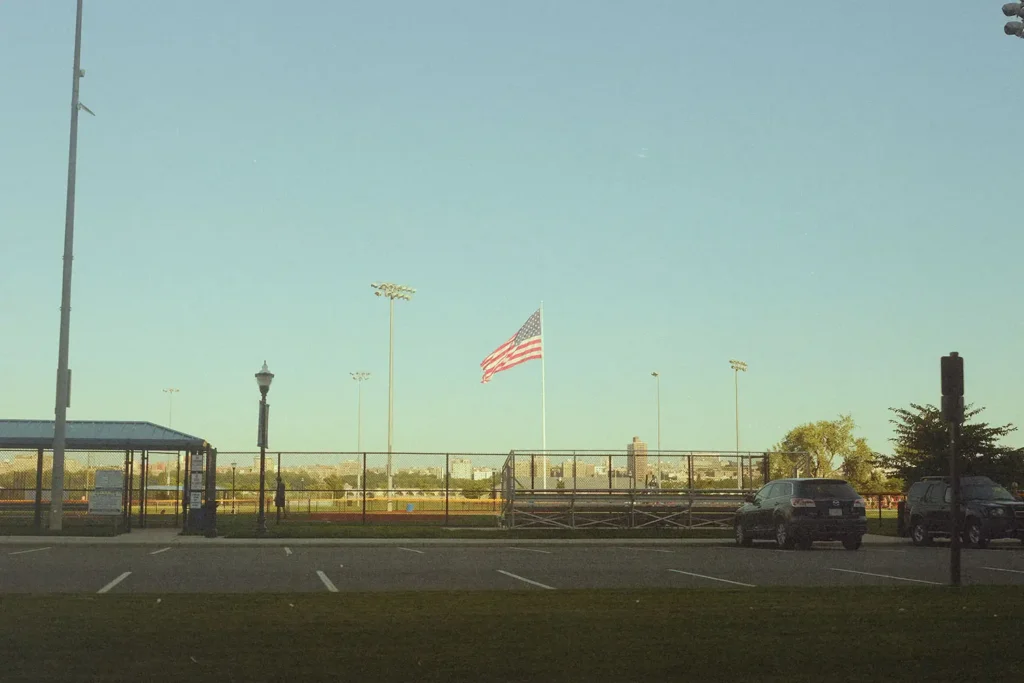 American flag flying in a ball field next to a parking lot