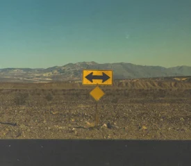 Yellow road sign with arrows pointing left and right in a barren desert landscape with distant mountains under a hazy sky