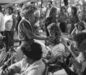 A crowded scene where Gloria Copeland places her hand gently on another person’s head while others around her sit or raise their hands