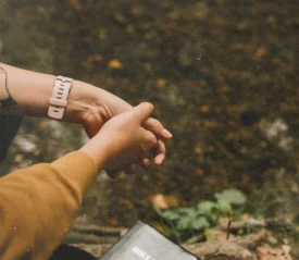 Hands clasped in prayer beside a closed Bible near a quiet stream in a natural setting