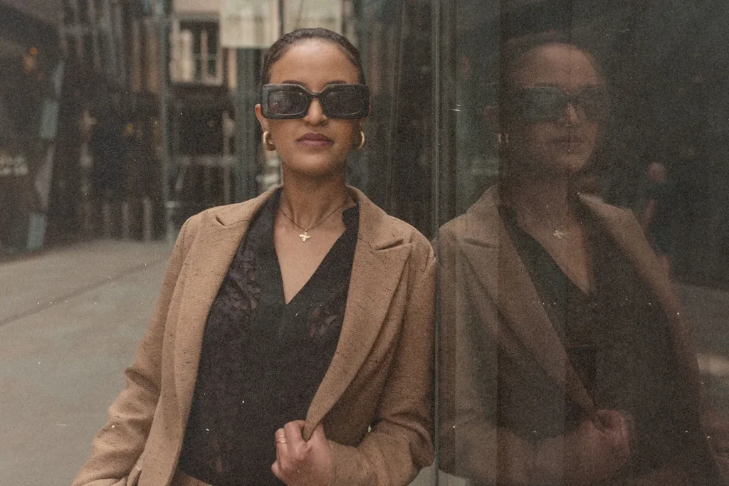 A person wearing a tan coat leans casually against a reflective glass wall in an urban setting