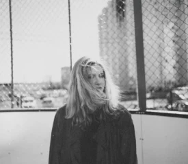 A person with long hair stands outdoors in front of a chain‑link fence, with sunlight shining through the background