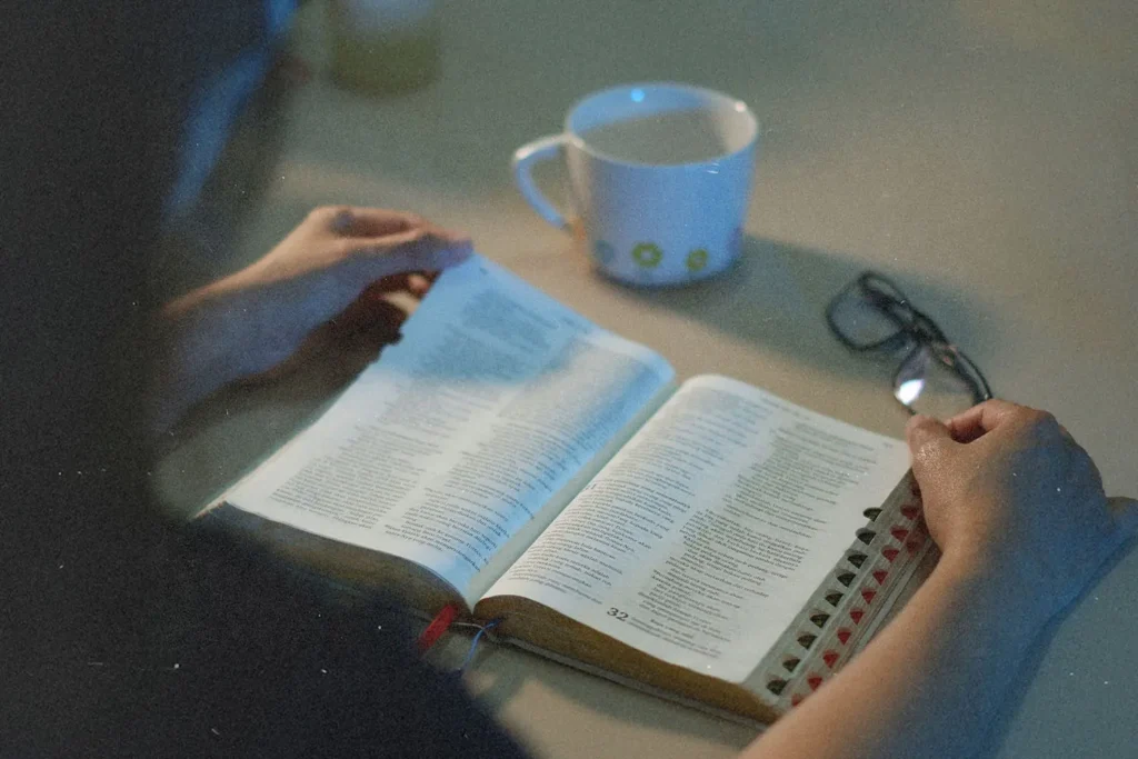 Person reading an open Bible at a table with a coffee cup and a pair of glasses nearby in soft, low light