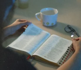 Person reading an open Bible at a table with a coffee cup and a pair of glasses nearby in soft, low light