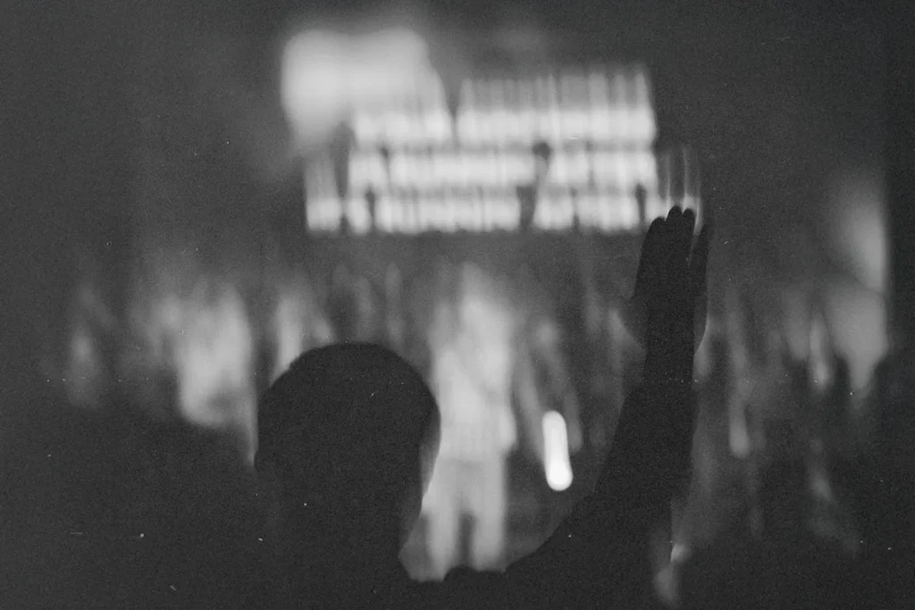 Silhouetted person raising one hand in front of a brightly lit stage with indistinct figures and text in the background