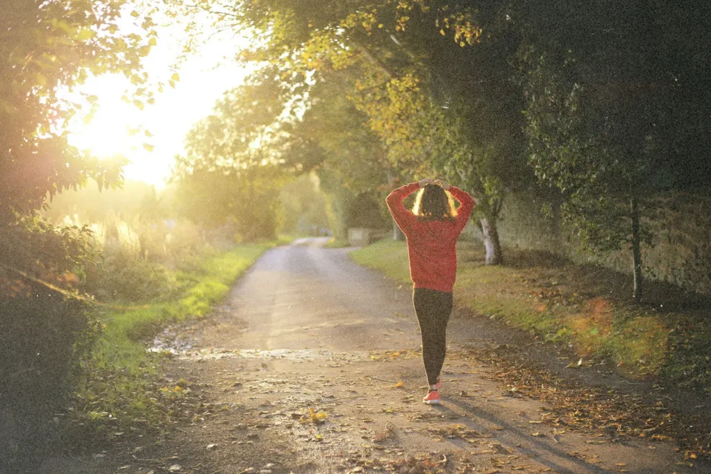 Person walking down a sunlit tree‑lined path at sunrise, wearing a red sweater and stretching with arms raised overhead
