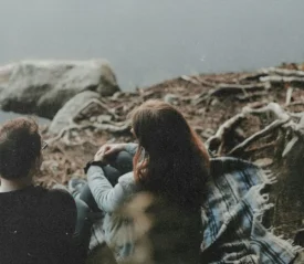 Two people sit on a blanket beside a calm lake, surrounded by tree roots and rocks along the shoreline