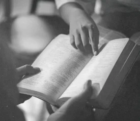Two people read an open Bible together, one person pointing to a passage on the page