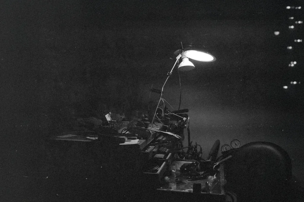 A dimly lit workshop scene shows a single overhead lamp illuminating a cluttered workbench filled with tools and machinery