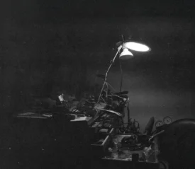 A dimly lit workshop scene shows a single overhead lamp illuminating a cluttered workbench filled with tools and machinery