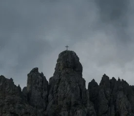 A cross stands atop a rugged mountain peak under a dark, overcast sky