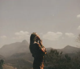 Person standing outdoors on a sunny day, holding a phone while overlooking distant mountains and a hazy landscape