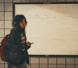 Person walking through a tiled subway station carrying a backpack and holding a phone, passing a large blank advertisement board with scribbles on it