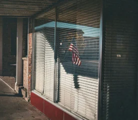 Window with open blinds and an American flag in the the glass reflection