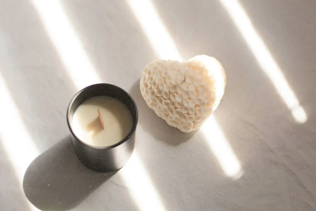 A black candle jar with a wooden wick sits beside a textured heart‑shaped object, both illuminated by bright streaks of sunlight