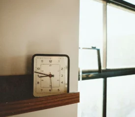 Square analog clock sitting on a wooden shelf next to a large sunlit window with metal frames
