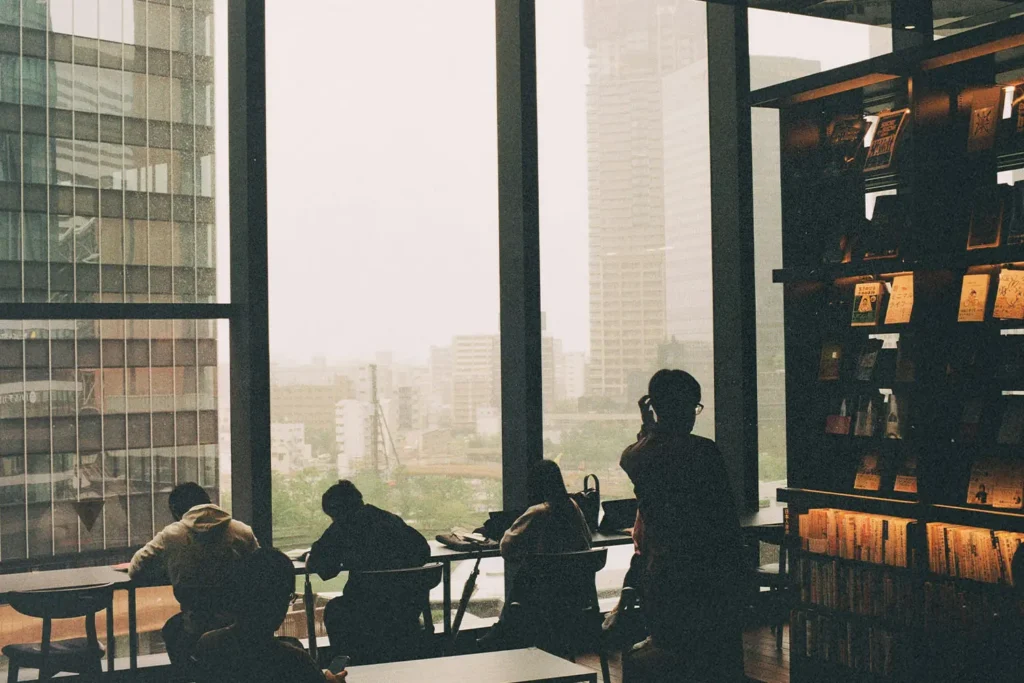 Several people sit and study near large floor‑to‑ceiling windows, overlooking a hazy cityscape from inside a modern library or café
