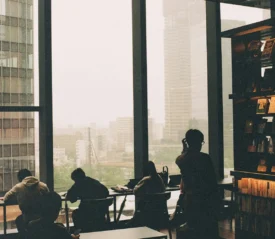 Several people sit and study near large floor‑to‑ceiling windows, overlooking a hazy cityscape from inside a modern library or café