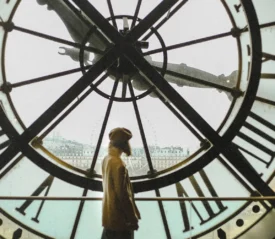 Person stands silhouetted inside a large clock tower, looking out through the transparent clock face toward a distant cityscape