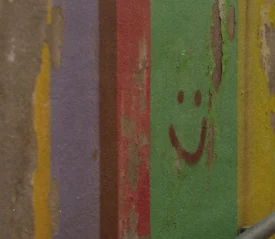 Peeling painted wall with vertical stripes of faded colors, including green, red, purple, yellow, and blue, featuring a simple smiley face drawn on the green section