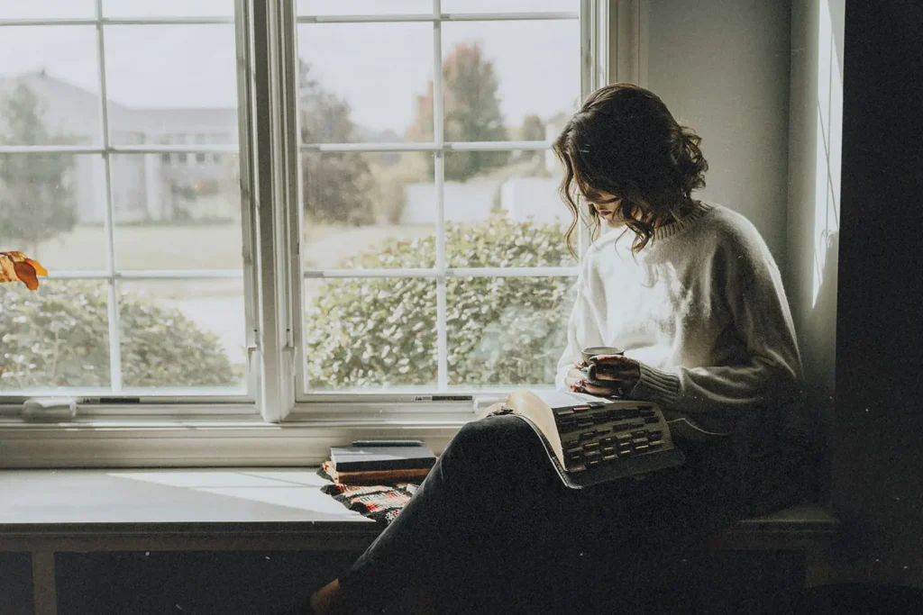 Woman sitting on a window seal studying her Bible with coffee cup in hand