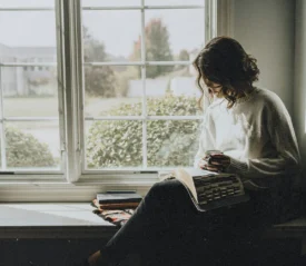 Woman sitting on a window seal studying her Bible with coffee cup in hand