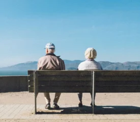 Two people sit on a wooden bench overlooking a bright blue sky and distant mountains