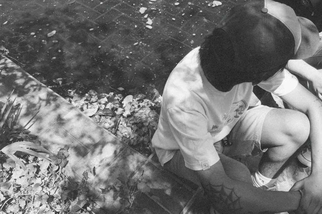 Black‑and‑white photo of a person sitting on outdoor steps in dappled sunlight, with fallen leaves scattered around