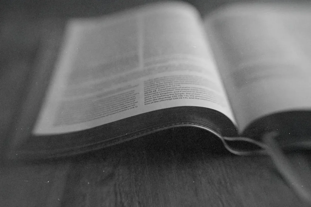 Open Bible lying on a wooden surface, with the pages softly curved and text slightly out of focus in a black‑and‑white image