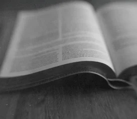 Open Bible lying on a wooden surface, with the pages softly curved and text slightly out of focus in a black‑and‑white image