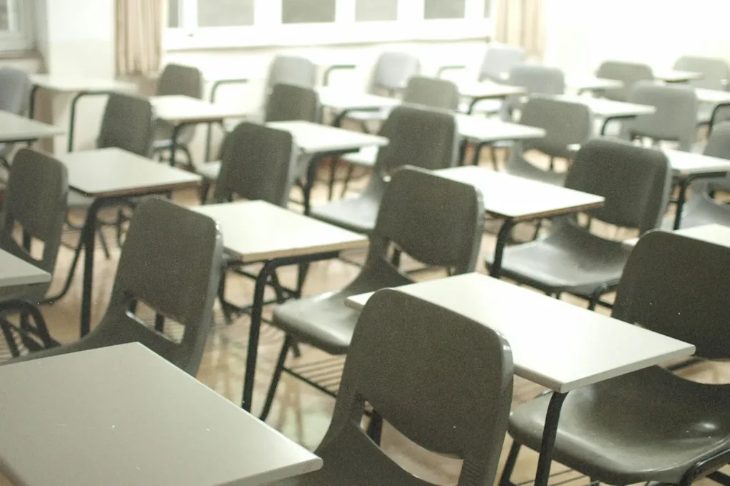 Rows of empty desks and chairs fill a bright classroom, with sunlight coming through large windows at the back of the room