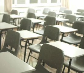 Rows of empty desks and chairs fill a bright classroom, with sunlight coming through large windows at the back of the room
