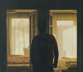 Silhouetted figure standing in a dimly lit doorway between two softly lit bedrooms with sheer curtains