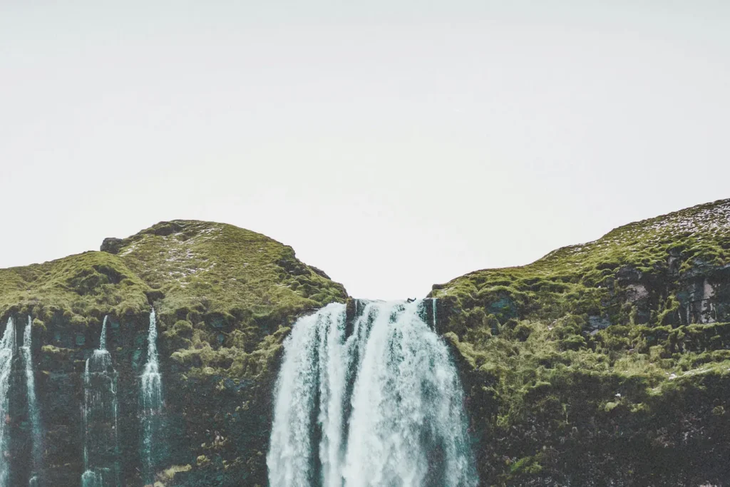 Wide waterfall cascading over a grassy cliff into a rocky valley under a bright, overcast sky