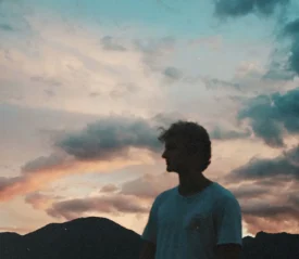 A person stands outdoors at sunset with mountains silhouetted behind them and colorful clouds filling the sky