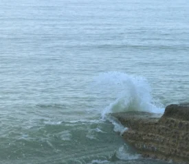 A person stands near the edge of a concrete ledge as a wave crashes against it, sending water upward
