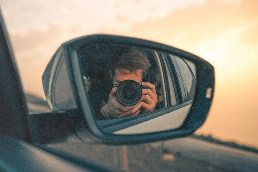 Photographer holding a camera reflected in a car’s side‑view mirror during a warm sunset drive