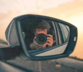 Photographer holding a camera reflected in a car’s side‑view mirror during a warm sunset drive