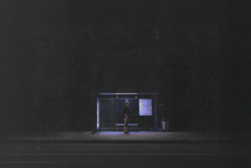 Single person stands under the dim blue light of a bus stop shelter at night, surrounded by darkness