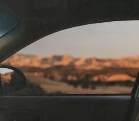 A view from inside a car looking out through the side window at a distant landscape, with sunlit hills and open countryside appearing softly out of focus