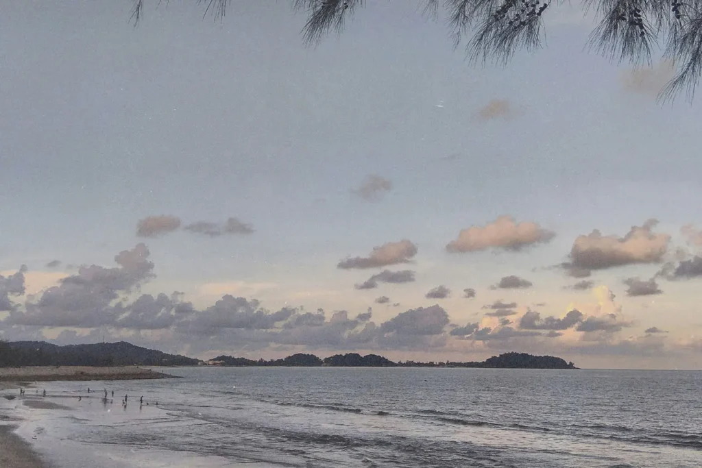 A calm beach scene at dusk with soft waves, scattered clouds, and distant tree‑covered hills across the shoreline