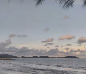 A calm beach scene at dusk with soft waves, scattered clouds, and distant tree‑covered hills across the shoreline