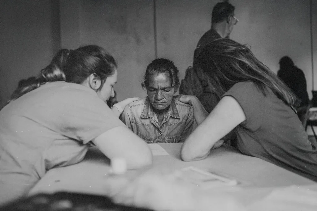 Three people sitting around a table in a quiet room, leaning in together in a moment of prayer
