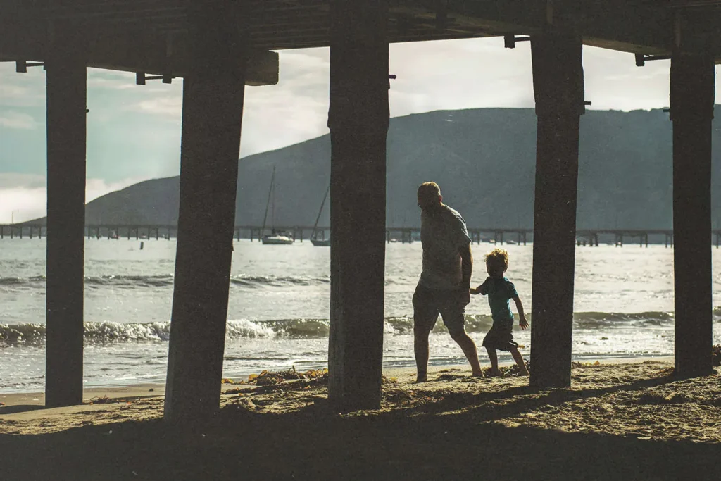 dad and son walking the beach under a pier