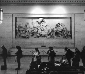 People standing and waiting inside a large hall with a detailed wall sculpture depicting historical figures, lit by tall hanging lamps