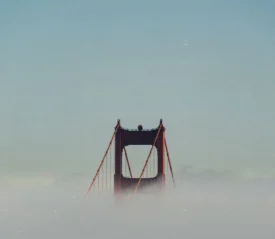 Top of a suspension bridge emerging through thick fog beneath a clear sky