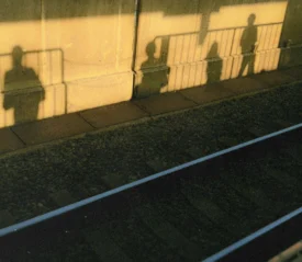 Long shadows of people standing on a train platform stretch across the wall and onto the ground in warm golden sunlight