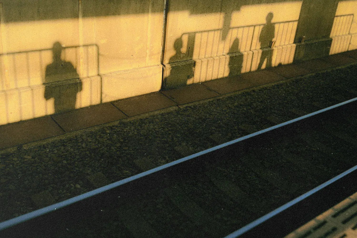 Long shadows of people standing on a train platform stretch across the wall and onto the ground in warm golden sunlight